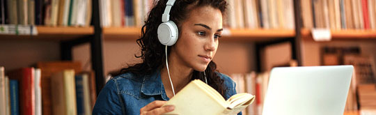 A person wearing headphones reads a book while using a laptop in a library.